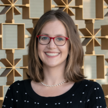 A smiling woman with shoulder-length brown hair and red glasses stands in front of a decorative geometric metal wall, wearing a black patterned top and a pearl necklace.
