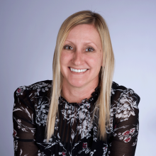 Amy headshot - caucasian woman with medium length blonde hair and light brown eyes wearing a black shirt with a white floral print