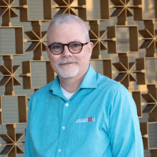 A man with short gray hair and glasses smiles gently while wearing a turquoise Amarillo button-down shirt, standing in front of a patterned metal wall with warm, geometric shapes.