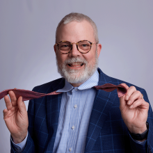 Sherman Headshot - Caucasian male with light gray hair and short beard playfully holding red untied tie open in a royal blue suit coat and light blue shirt