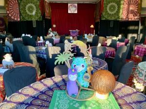 Colorful, bohemian-style table settings inside The Lobby Lounge at The Harbor Center, with patterned tablecloths, whimsical centerpieces, llama stuffies and a small stage framed by red curtains in the background.