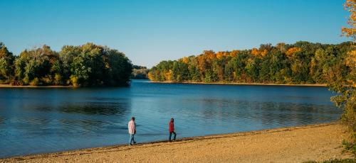 Image of two people strolling along the lake shore in fall under a bright blue sky