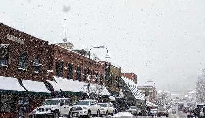 A row of brick buildings alongside a road have snow covered awnings and roofs. There are cars lined up on the road a snowflakes visible in the sky.