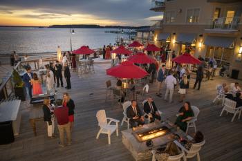 A group of people are shown attending a conference event at the Lighthouse Cove Event Center in Dewey Beach, Delaware as the sun sets in the background. Some people in the photo are standing while others are seated around fire pits or sitting under tables at red umbrellas.