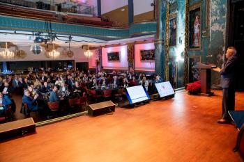 A man stands at a podium on a stage speaking to a group of people seated in front of the stage during a conference held at The Queen in Wilmington, Delaware.