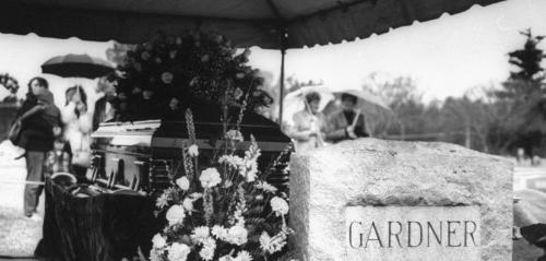 Black and white photo of Ava's gravestone with coffin and flowers and mourners under umbrellas in the back