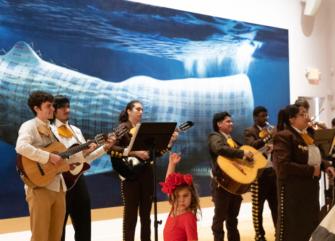 A group of musicians playing guitars and singing with a child in front of a large blue wall.