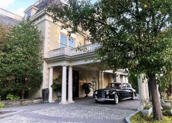 A large, beige building with columns and a balcony, with a black vintage car parked in front.