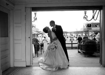 A black and white photo of a couple dancing in the doorway of a building with outdoor seating in the background.