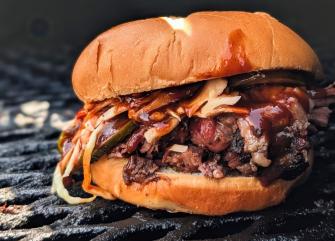 A grilled burger with barbecue sauce, coleslaw, pickles, and a beef patty on a bun.