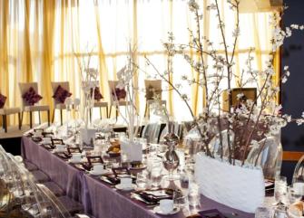 A dining room with a long table set for a meal, featuring purple tablecloth, white flowers, and a window with sheer curtains.