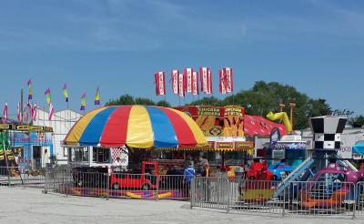 A colourful carnival midway with food trucks, children’s rides, striped canopies, and flags on a sunny day.