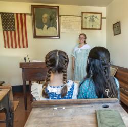 Picture of a colonial era school room with a teacher at the front and two pupils sitting at a desk at the Warrior Run Fort Freeland Heritage Days Festival. A big American flag hangs on the wall next to a picture of George Washington.