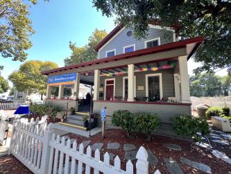 Front view of Casa de Cultura y Arte, a restored Craftsman bungalow with red roof and trim, wraparound porch, and white picket fence in Gilroy