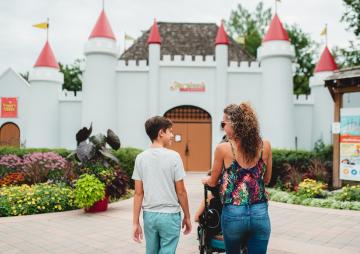 A woman and child walk toward the castle-style entrance of Storybook Gardens, surrounded by colourful flower beds.