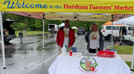 A male (left) and female (female) wearing white t-shirts with the Horsham Farmers Market graphic logo on the front and jackets standing in front of a table with a table cloth on top reading Horsham Farmers Market. They are posed under the yellow tent that reads Welcome to the Horsham Farmers' Market. The wet parking lot and loading trucks appear in the background.