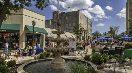 Suburban Square located in Ardmore, PA. The fountain is located in the middle of the shopping district. In the background is the storefront of Barbour. Vendors are seated at tables talking to customers. Many individuals are seen walking around enjoying the warm weather or seated outside for dining at the tables with blue umbrellas.