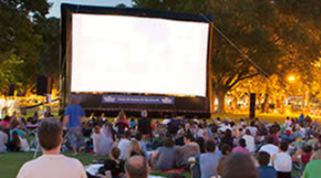 A large group of movie goers wait for the movie to begin on the inflatable screen in the park. Many attendees are sitting in lawn chairs or laying on blankets in the grass. The trees are lit by the park lights in the background.