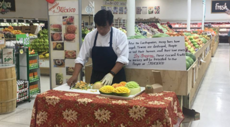 A man wearing a white chef's shirt and black apron stand behind a table with a red and yellow floral tablecloth while cutting and sculpting produce. Rows of various bins of produce are seen in the background.