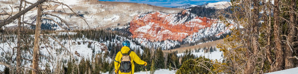 Skier dressed in bright yellow coat looks out at red rock formations dusted in snow at Brian Head Resort, UT.