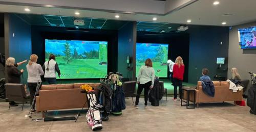 Women at indoor golf facility - with couches and clubs and two screens of virtual golf courses