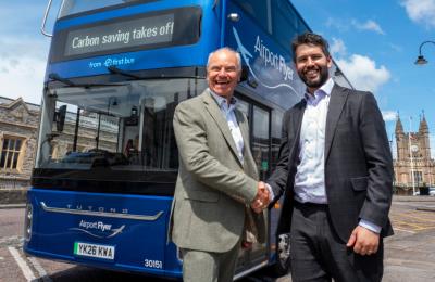 Two men shaking hands in front of a dark blue electric bus parked outside Bristol Temple Meads railway station.