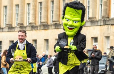A man dressed in Regency costume and a man dressed in a Frankenstein costume running along Bath's Royal Crescent while holding trays containing a champagne flute and bun.