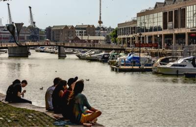 People sitting on the Bristol Harbourside with Pero's Bridge and the cranes outside M Shed in the background