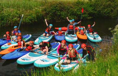 A large group of young people on paddleboards and kayaks is rafted up in a group on the River Yeo, smiling and waving in bright sunshine.