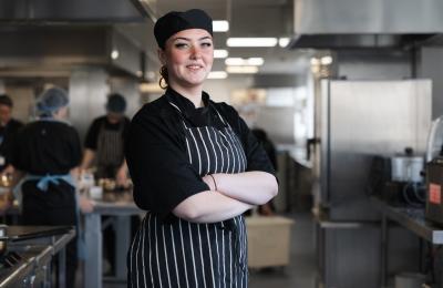 A chef standing with their arms folded and smiling in the centre of a commercial kitchen.