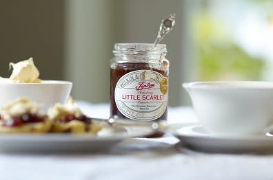 An Open pot of Tiptree Little Scarlet Jam sits, opened, beside scones, cream and a cup of tea on a pristine tablecloth.