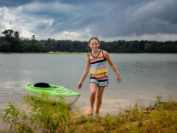 A little girl pulls a kayak out of the lake while smiling at the camera