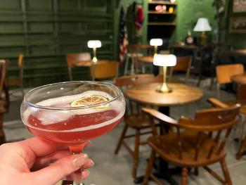 A hand holds a pink cocktail up in a glass in front of the camera, the interior of the bar can be seen in the background.