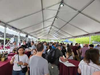 A large crowd of people samples food and wine under a tent.