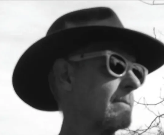 A black and white outdoor photo of a man looking off to the right. He is wearing a stylish wide-brimmed hat and plastic sunglasses.