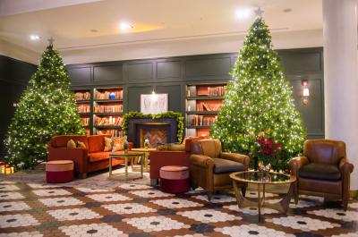 x2 giant Christmas trees bookending comfy seating at the Clayton Hotel Cambridge. There are bookcases in the background with an open fireplace in the middle that has green tinsel on its surround.