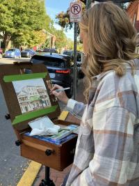 Woman standing at an easel painting outside with a street view and trees in the background as part of the Selinsgrove Plein Air Painting Competition