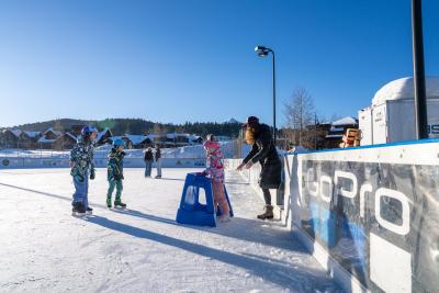 Children in colorful winter gear practice skating on an outdoor ice rink, with a snowy landscape and mountains in the background. A coach assists them.