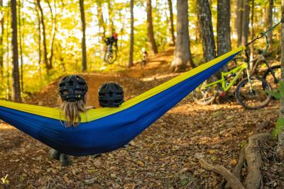 A child and an adult sit in a hammock overlooking mountain bike trails with mountain bikers.