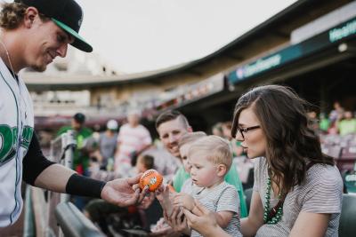 A Dayton Dragons player handing a baseball to a toddler while the mother looks on.
