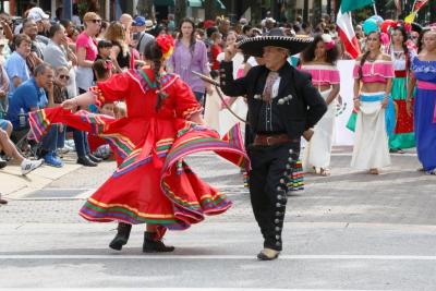 Man and woman dressed in vibrant traditional Mexican attire dance joyfully in a street parade during Fayetteville’s International Folk Festival, surrounded by colorful costumed performers and a cheering crowd.