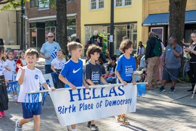 Children dressed in traditional and modern Greek-themed outfits proudly march in a parade, holding a banner that reads "HELLAS – Birth Place of Democracy," during the Fayetteville, NC Greek Festival. Spectators line the sidewalk, watching and cheering.