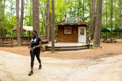 Smiling woman walks solo past the rustic ZipQuest Adventure Center, surrounded by tall pine trees and wooden benches in a peaceful forest setting.