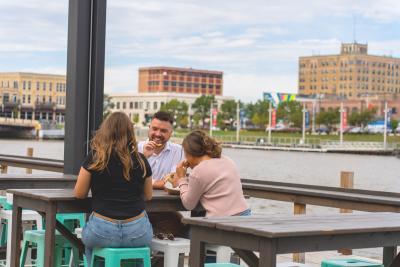 people eating outside at The Wharf with view of the Manitowoc River and buildings