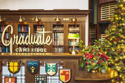 Graduate by Hilton hotel in Cambridge front reception. A glass bookcase is the background with wooden plaques of individual college flags in the foreground. To the right is a mini bushy Christmas tree on a side table with a much bigger christmas tree to the right with warm yellow lights