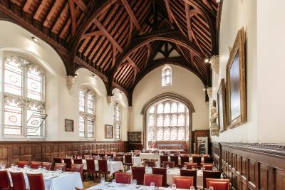Ridley Hall Cambridge dining hall - wooden beamed ceiling with large arch stained glass windows and light cream coloured walls. Tables and chairs are set in sprigs with white linen and silver tableware