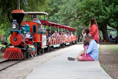 A young family sits on the sidewalk at Landa Park to watch the Landa Park train pass by.