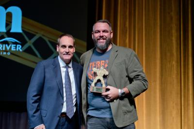 Chef Brad Checci, wearing a blue Terra Madre Americas tee beneath a green blazer, accepts the State of Tourism Hospitality Hero award depicting a superhero