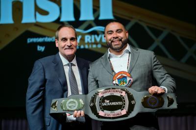 Wilton Rancheria Chairman Jesus Tarango, wearing a gray suit and the great seal of the Wilton Rancheria tribe, accepts the Tourism Champion award, a wrestling-style engraved belt