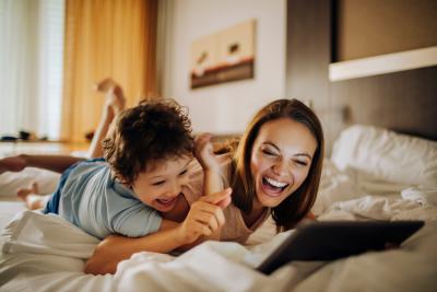 Woman and child laughing, watching tablet together on hotel bed